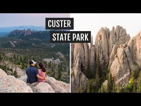 A couple sits on a rocky ledge at Custer State Park, overlooking a forest and distant hills. Nearby, rugged rock formations rise vertically amidst the trees.